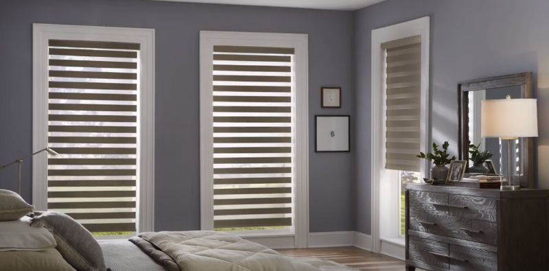 A bedroom with a bed , dresser , mirror and striped dual shades on the windows. Love is Blinds